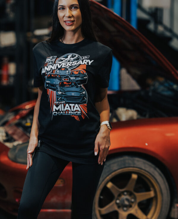 Woman wearing a Miata anniversary T-shirt at a car workshop, celebrating Mazda Miata's milestone with a vibrant design and automotive theme.
