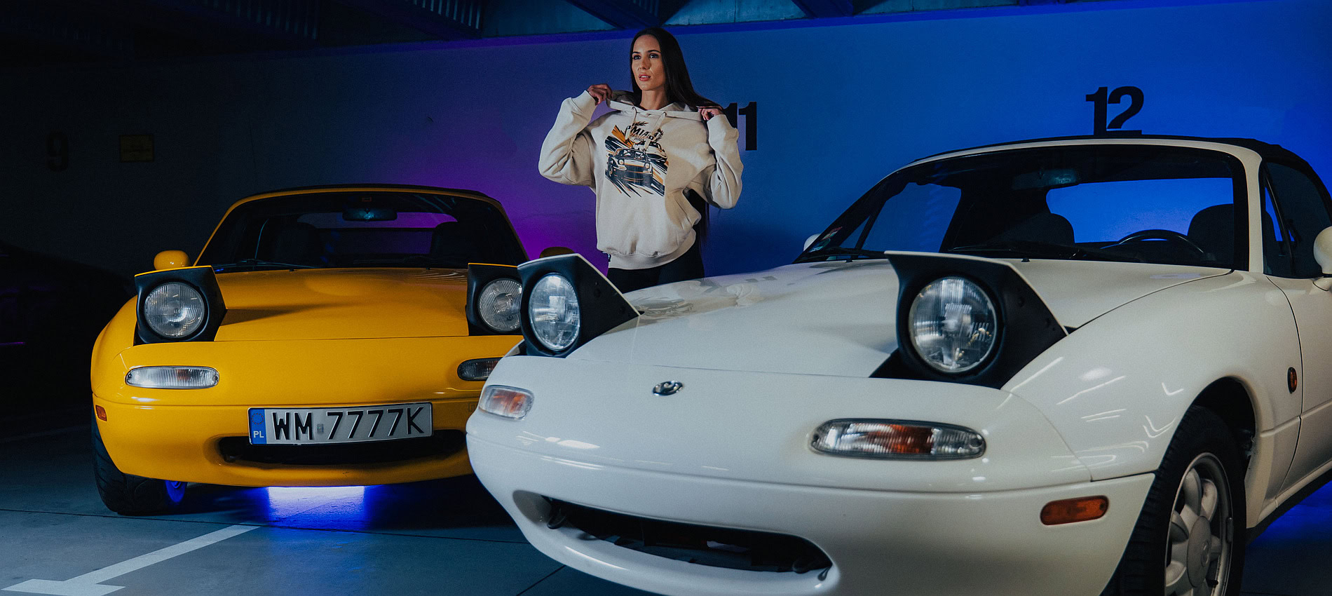 Woman standing between two classic Mazda Miatas in a showroom, celebrating the spirit of Miata with vibrant cars and a passionate car enthusiast vibe.