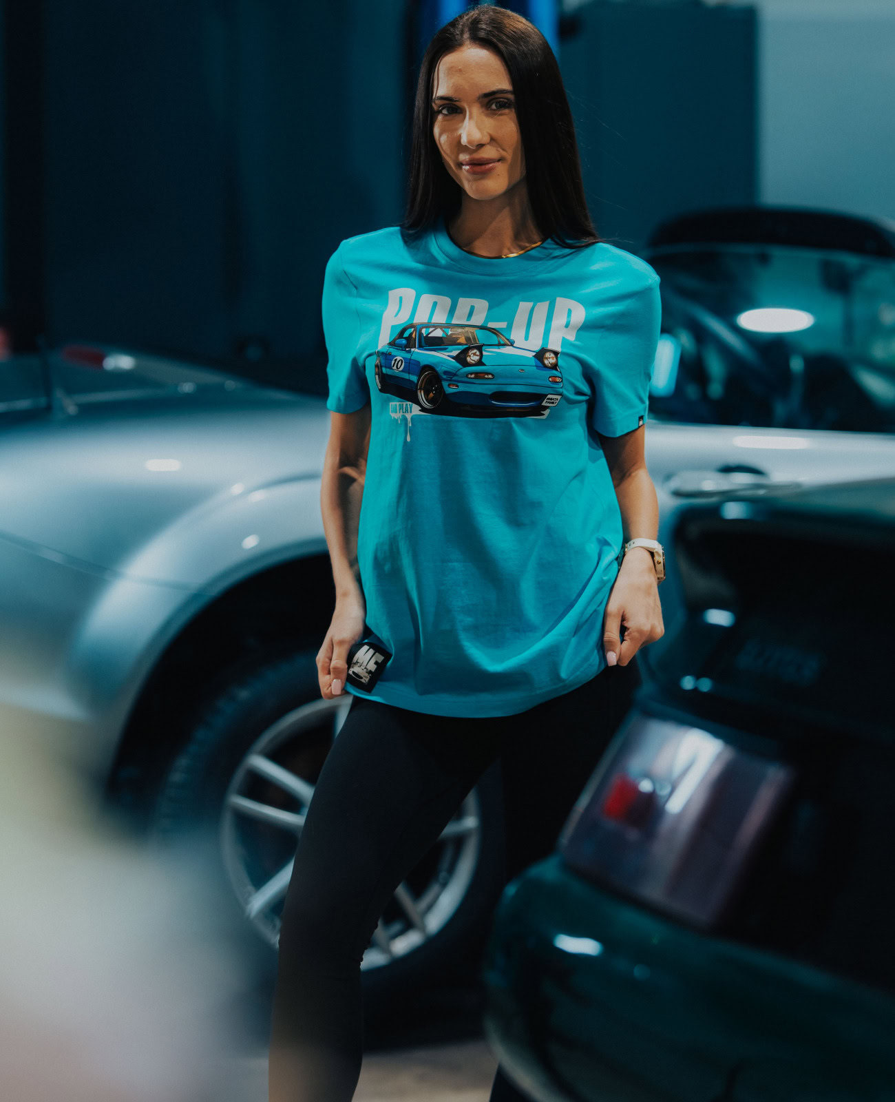 Woman wearing a blue Miata-themed T-shirt at a car show, surrounded by various Mazda Miata vehicles in a well-lit indoor setting.