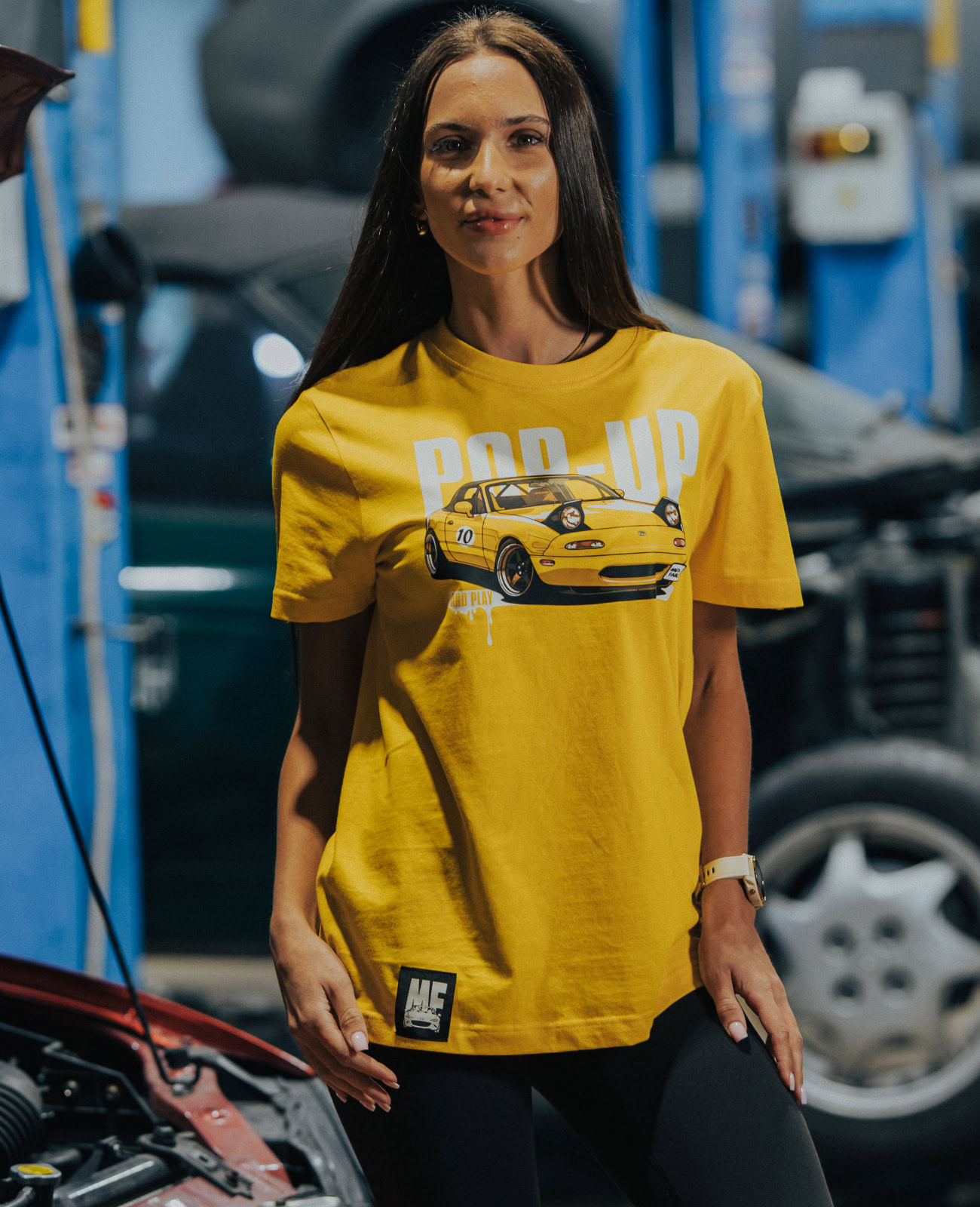 Enthusiastic woman wearing a yellow Mazda Miata T-shirt with "POP UP" text and car graphic, standing in a garage or auto shop environment.